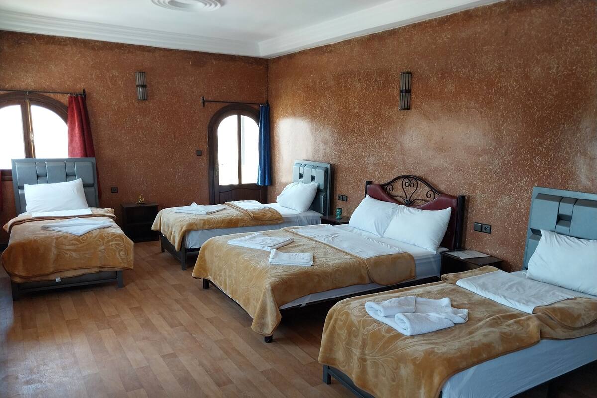 Quarto de hotel com quatro camas de solteiro, cada uma com colcha marrom e almofadas brancas. O quarto possui piso de madeira e parede marrom texturizada.