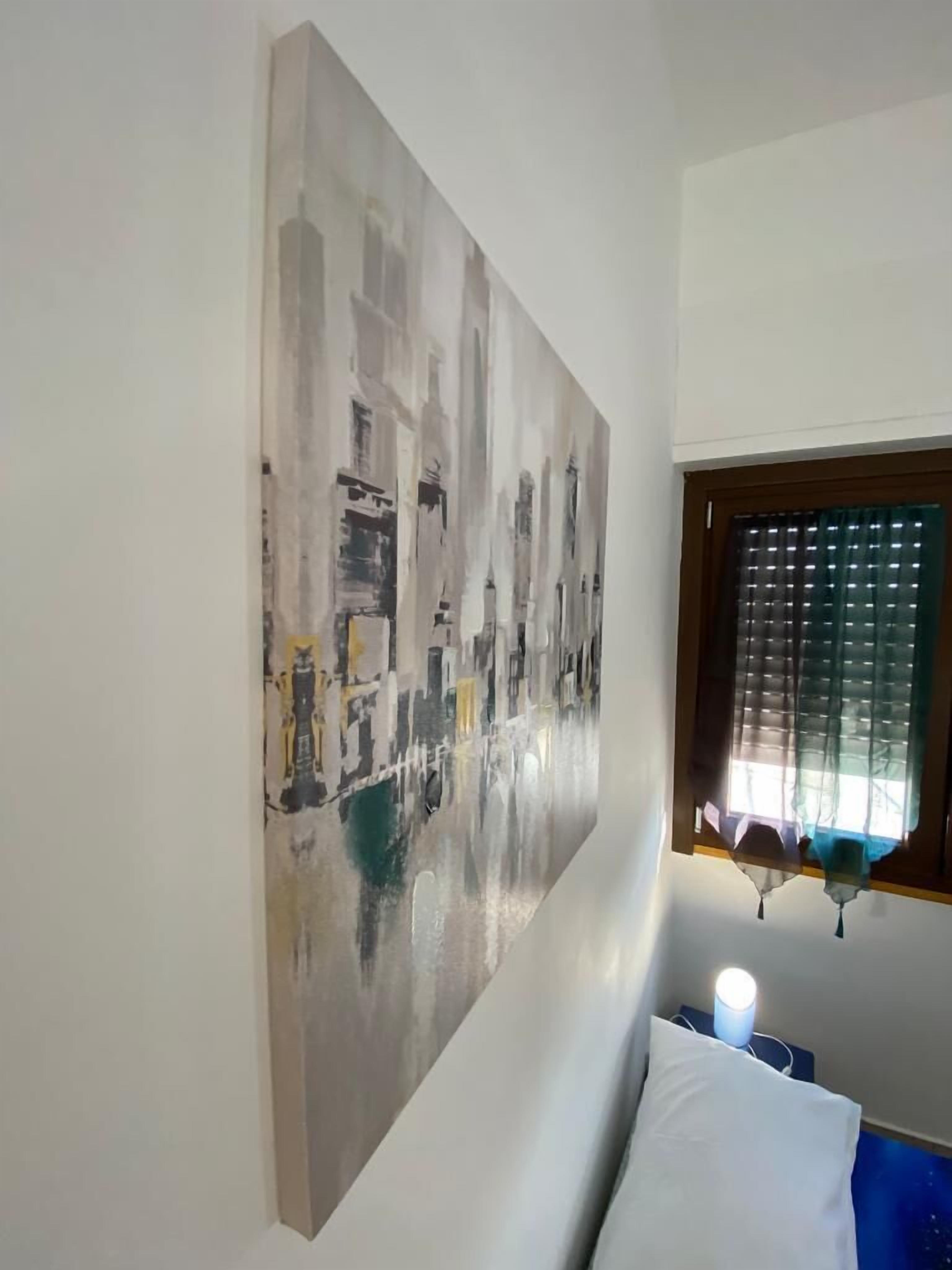 A bedroom with a large abstract painting on the wall, a bed with a white pillow, and a window with blinds.