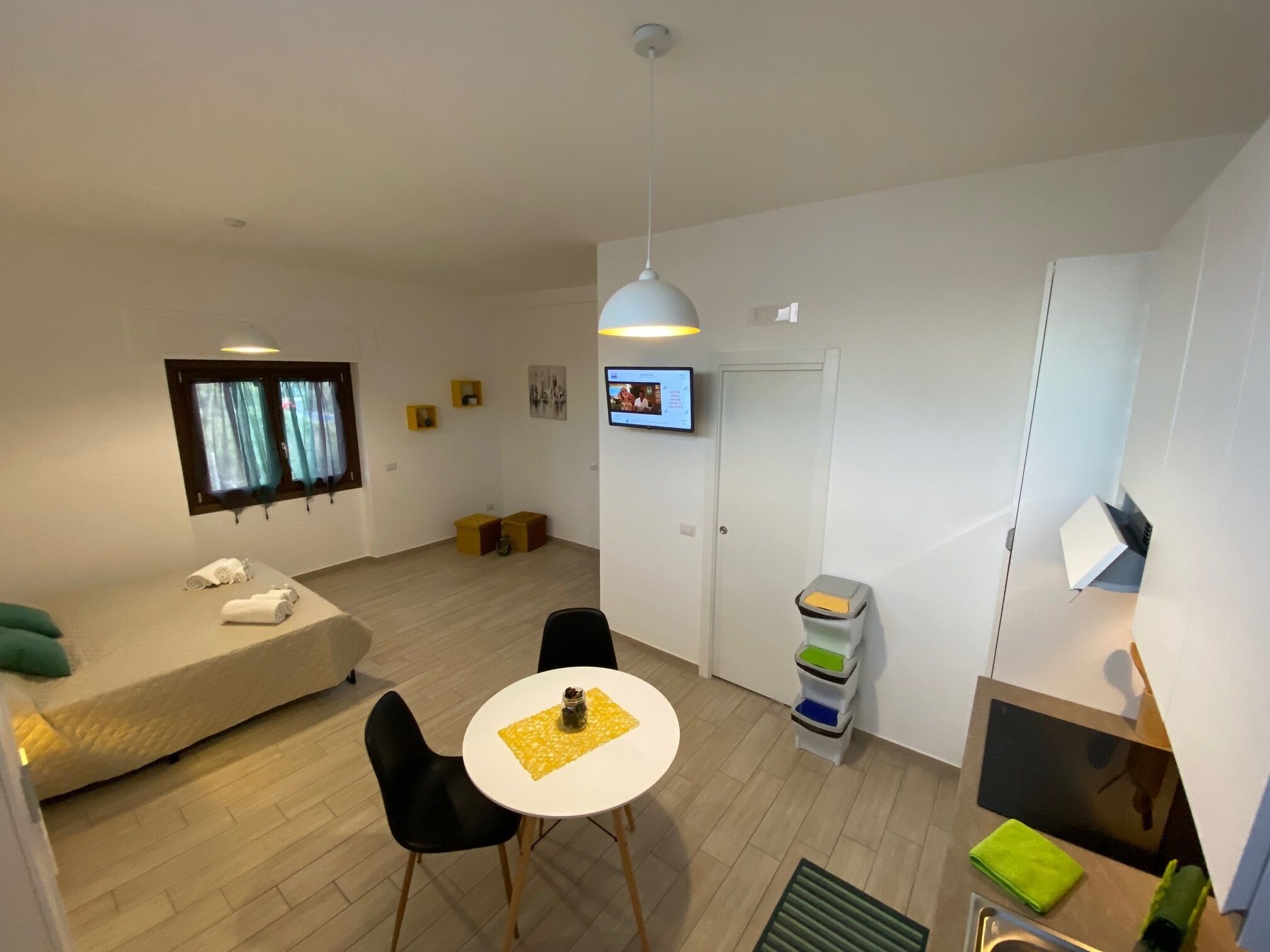 Ein modernes, gut beleuchtetes Zimmer mit einer Couch, einem Esstisch mit Stühlen, einem an der Wand befestigten Fernseher und einer kleinen Küchenzeile mit Spüle und Stauraum.