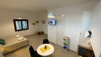 Ein modernes, gut beleuchtetes Zimmer mit einer Couch, einem Esstisch mit Stühlen, einem an der Wand befestigten Fernseher und einer kleinen Küchenzeile mit Spüle und Stauraum.