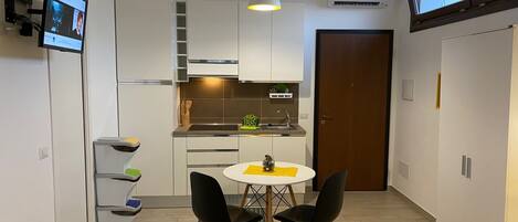 Una cocina compacta con armarios blancos, una mesa de comedor con dos sillas, una lámpara colgante y un televisor montado en la pared.