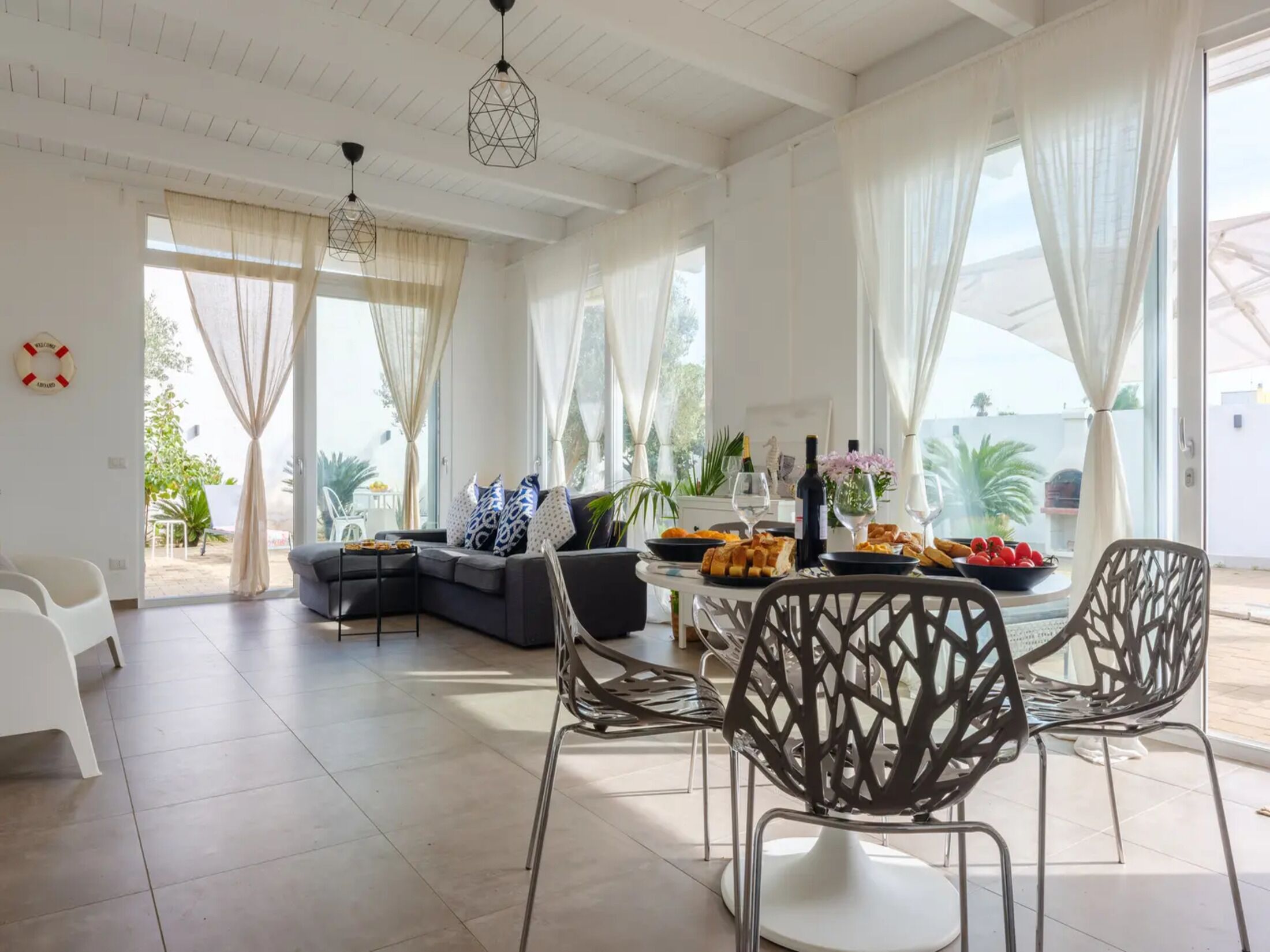 Un vaste espace de vie agrémenté d’un canapé moderne, d’une table à manger dressée avec de la nourriture et des boissons, et de grandes fenêtres ornées de rideaux légers.