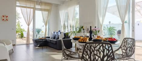 Un salón amplio con un sofá moderno, una mesa de comedor preparada con comida y bebidas, y ventanas grandes con cortinas translúcidas.