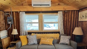 A cozy cabin interior with a grey sofa, yellow and patterned cushions, a lamp, and a window with a view of a car outside. - Canary Cottage: Lakeside, Boat Slip, Beach, Family Friendly (Ludington)