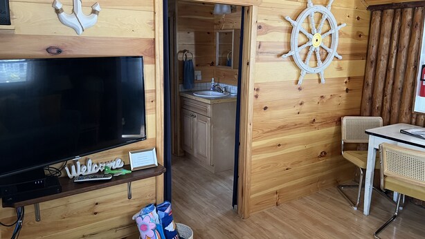 A wooden cabin interior with a television, a welcome sign, a ship's wheel, and a small dining area. - Canary Cottage: Lakeside, Boat Slip, Beach, Family Friendly (Ludington)