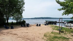 A sandy beach with several Adirondack chairs, a swing set, and a dock with boats. - Canary Cottage: Lakeside, Boat Slip, Beach, Family Friendly (Ludington)