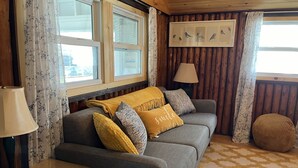 A cozy living room with a grey sofa, yellow throw pillows, a lamp, and a framed picture of birds on the wall. - Canary Cottage: Lakeside, Boat Slip, Beach, Family Friendly (Ludington)