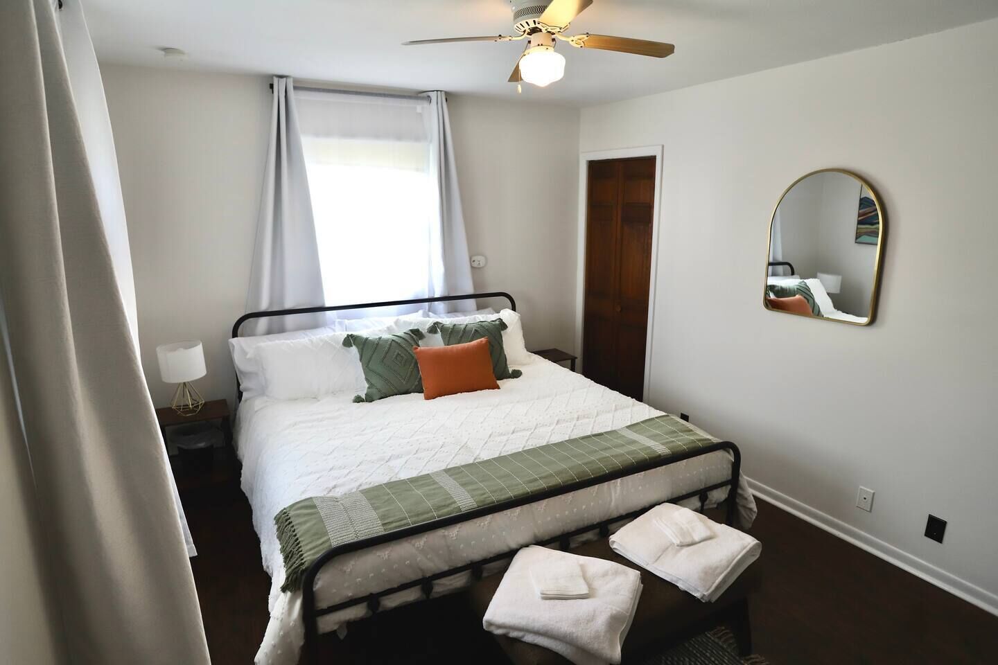 Un dormitorio con cama, ventilador de techo, ventana con cortinas, puerta de madera y espejo en la pared.