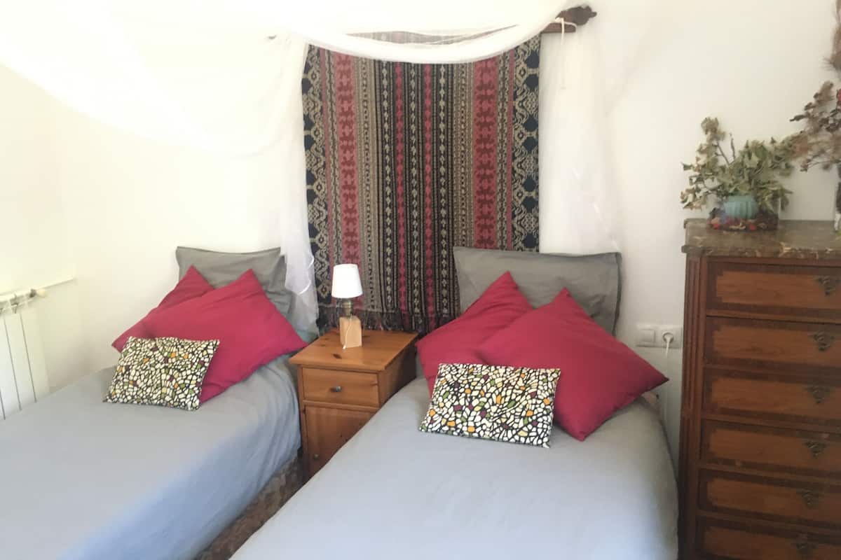 A room with two beds, each with a red pillow and a patterned pillow, a wooden nightstand with a lamp, and a patterned curtain.