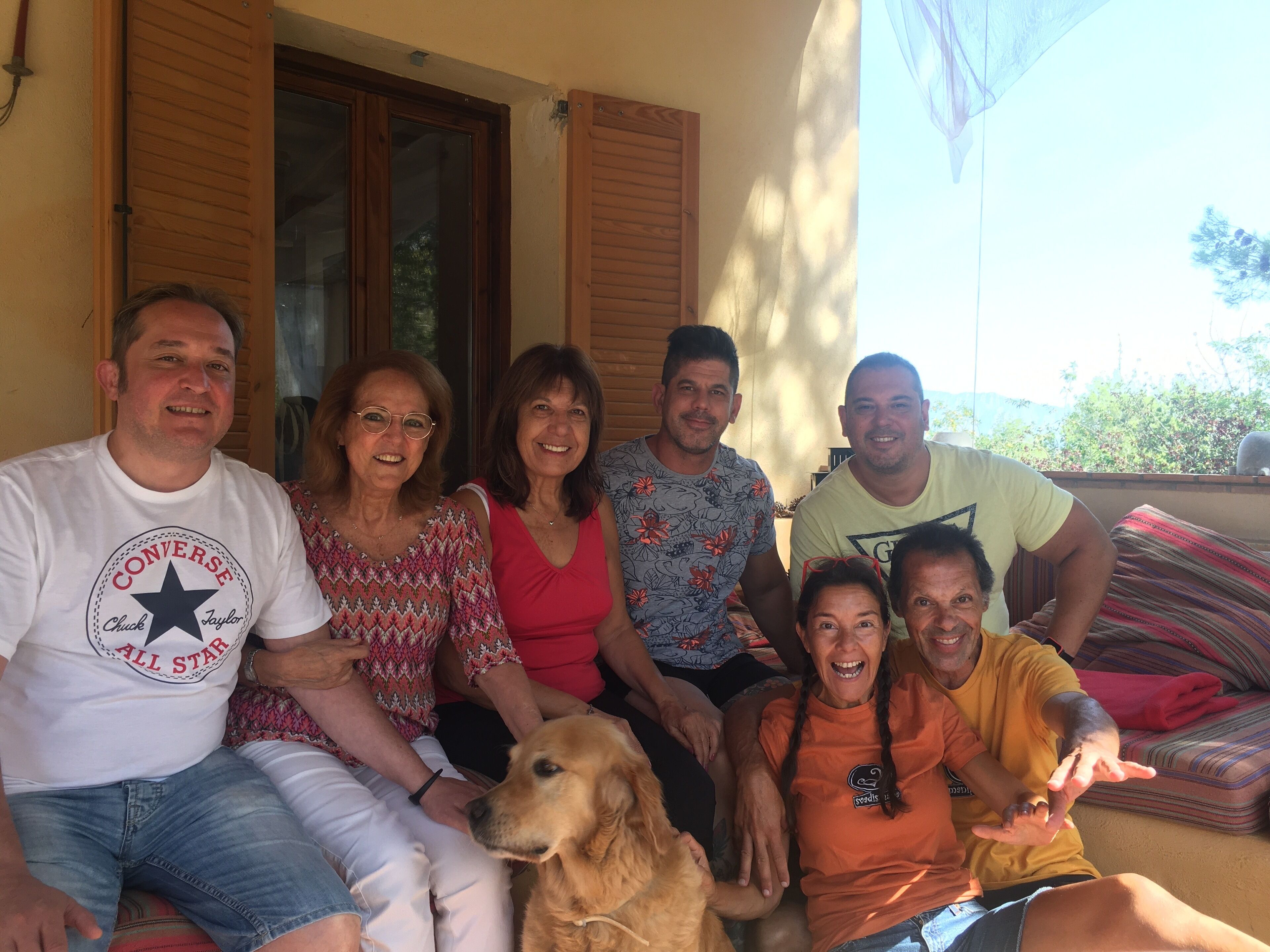 A group of people sitting on a porch with a dog, wooden shutters, and a view of greenery.