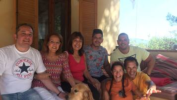 A group of people sitting on a porch with a dog, wooden shutters, and a view of greenery.