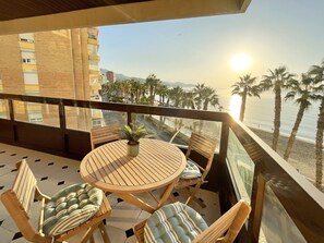 A balcony with a round wooden table and chairs, a potted plant, and a view of the beach and palm trees.