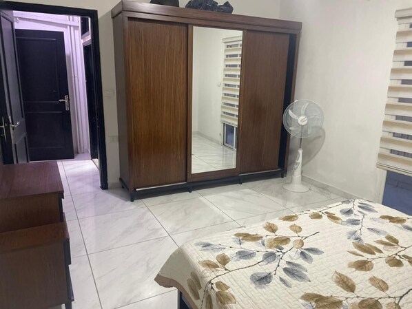 A bedroom with a wardrobe, a bed with a floral bedspread, a standing fan, and a tiled floor.