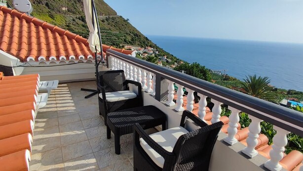 Ein Balkon mit Korbstühlen und einem Tisch, der Blick auf eine Küstenlandschaft mit Terrakottafliesen und einer weißen Brüstung bietet.
