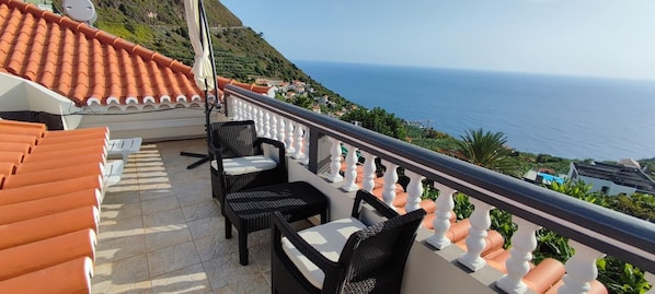 Ein Balkon mit Korbstühlen und einem Tisch, der Blick auf eine Küstenlandschaft mit Terrakottafliesen und einer weißen Brüstung bietet.