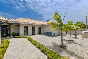 A building with a tiled roof, a stone pathway, and a small garden area with a bench and palm trees.
