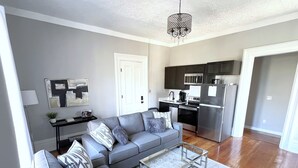 A modern living room with a grey sectional sofa, a glass coffee table, and a kitchen area with dark cabinets and stainless steel appliances.