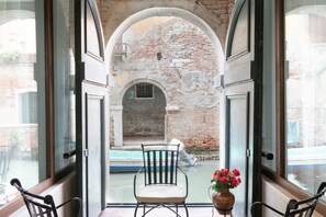 Une pièce dotée de grandes fenêtres, donnant sur une cour avec un mur en briques, comprenant une chaise et un vase rempli de fleurs.