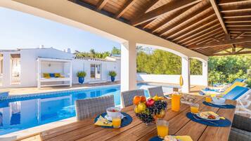 Un espace repas aménagé au bord de la piscine, avec une table dressée pour le petit-déjeuner, donnant sur une piscine et un bâtiment blanc doté d’un toit recouvert de tuiles.