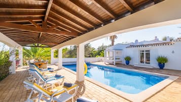 Un área de piscina cubierta al aire libre con sillones de descanso y vista a un edificio blanco.