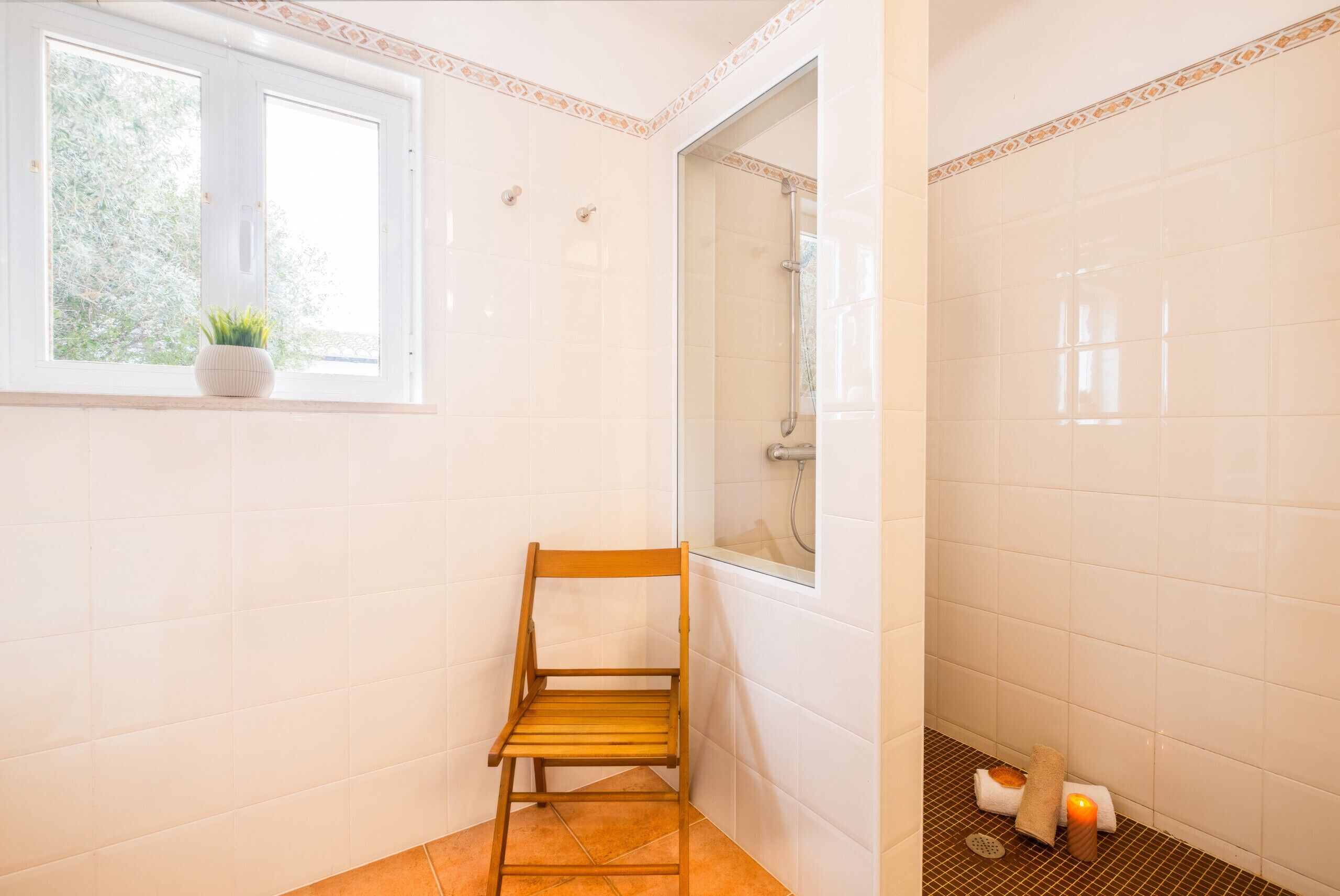 A bathroom with white tiles, a wooden chair, a window with a plant, and a shower area.