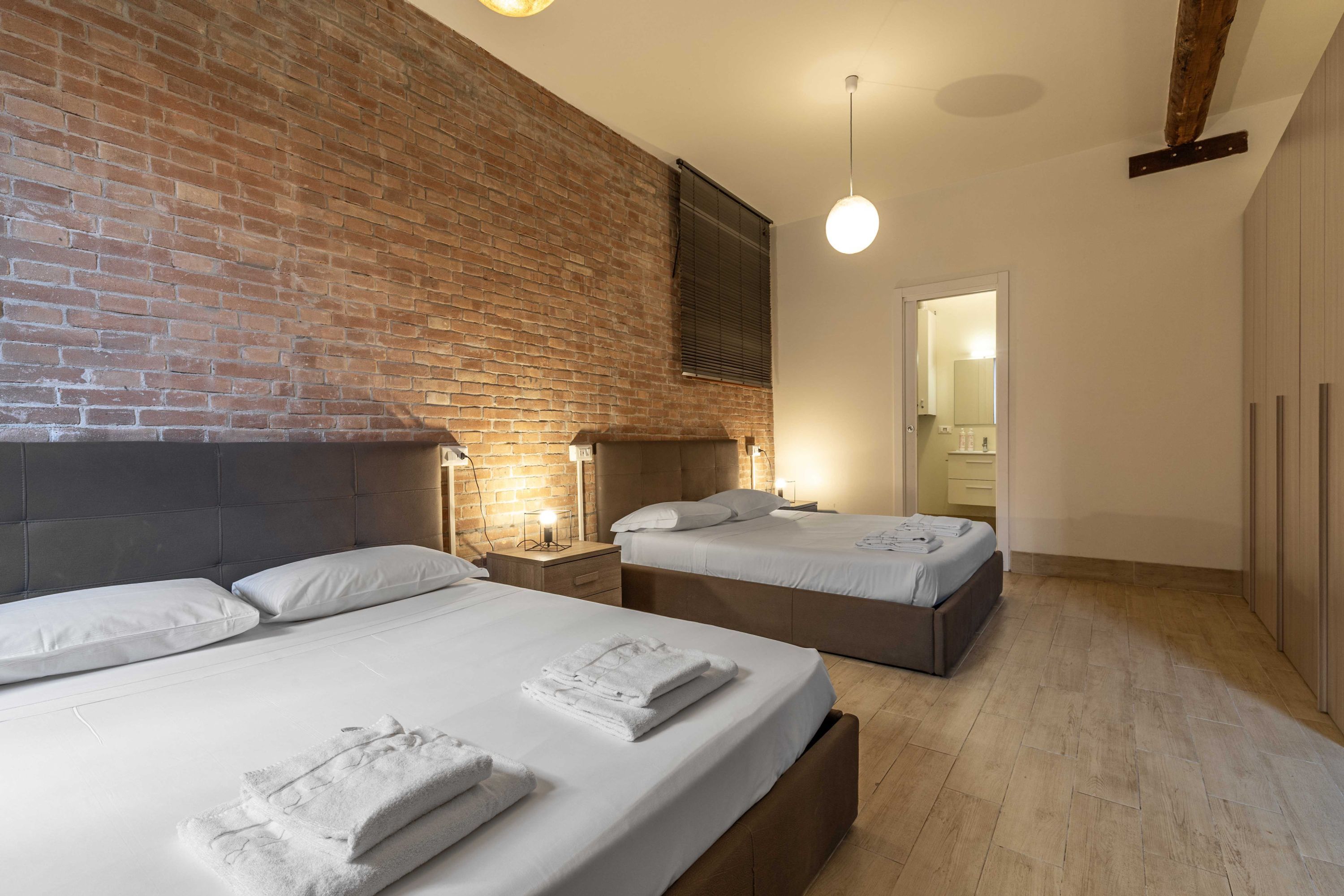 A hotel room with a brick wall, two beds, bedside lamps, and a wooden floor.