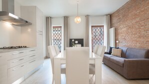 Une cuisine moderne avec des meubles blancs et une cuisinière à gaz, un coin repas avec une table et des chaises blanches, et un salon avec un canapé marron et un mur en briques.