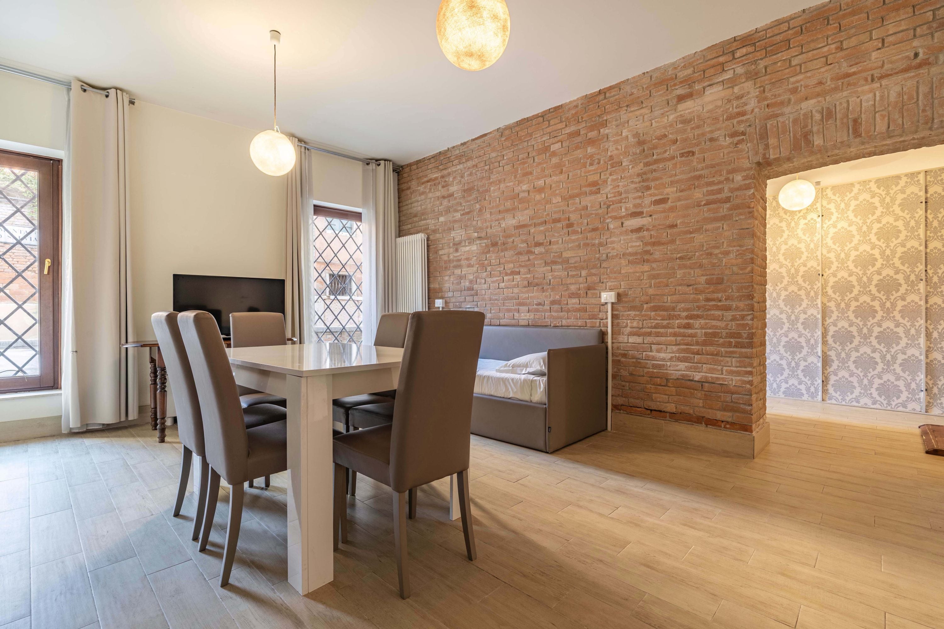 A modern room with a dining table, chairs, a sofa bed, a brick wall, and a window with a view.