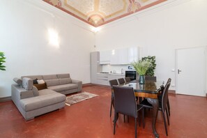 Un salon avec un canapé d’angle gris, une table à manger avec des chaises et un coin cuisine doté de meubles blancs et d’une porte blanche.
