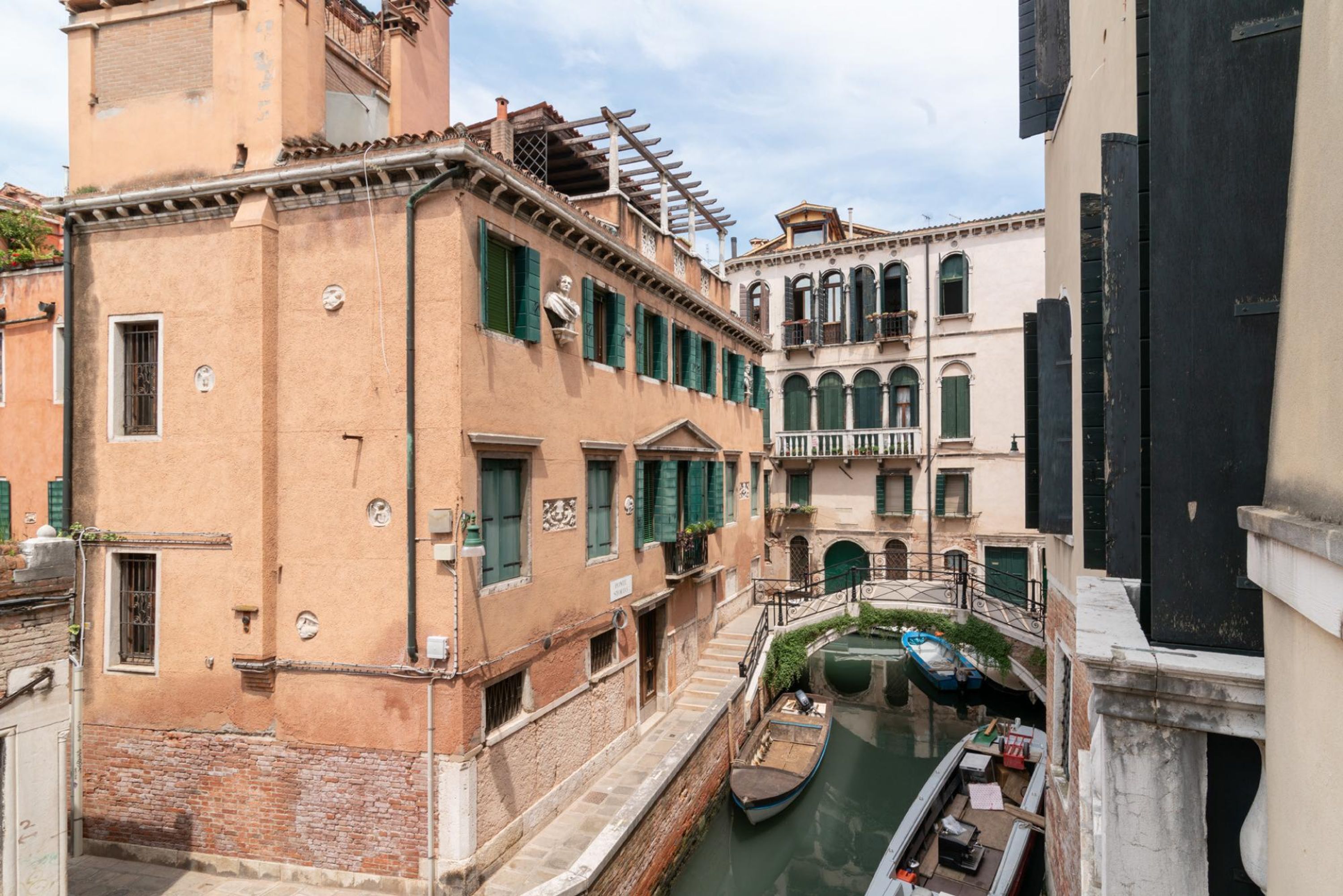 A narrow canal with a boat docked on the right, surrounded by historic buildings with balconies and green shutters.