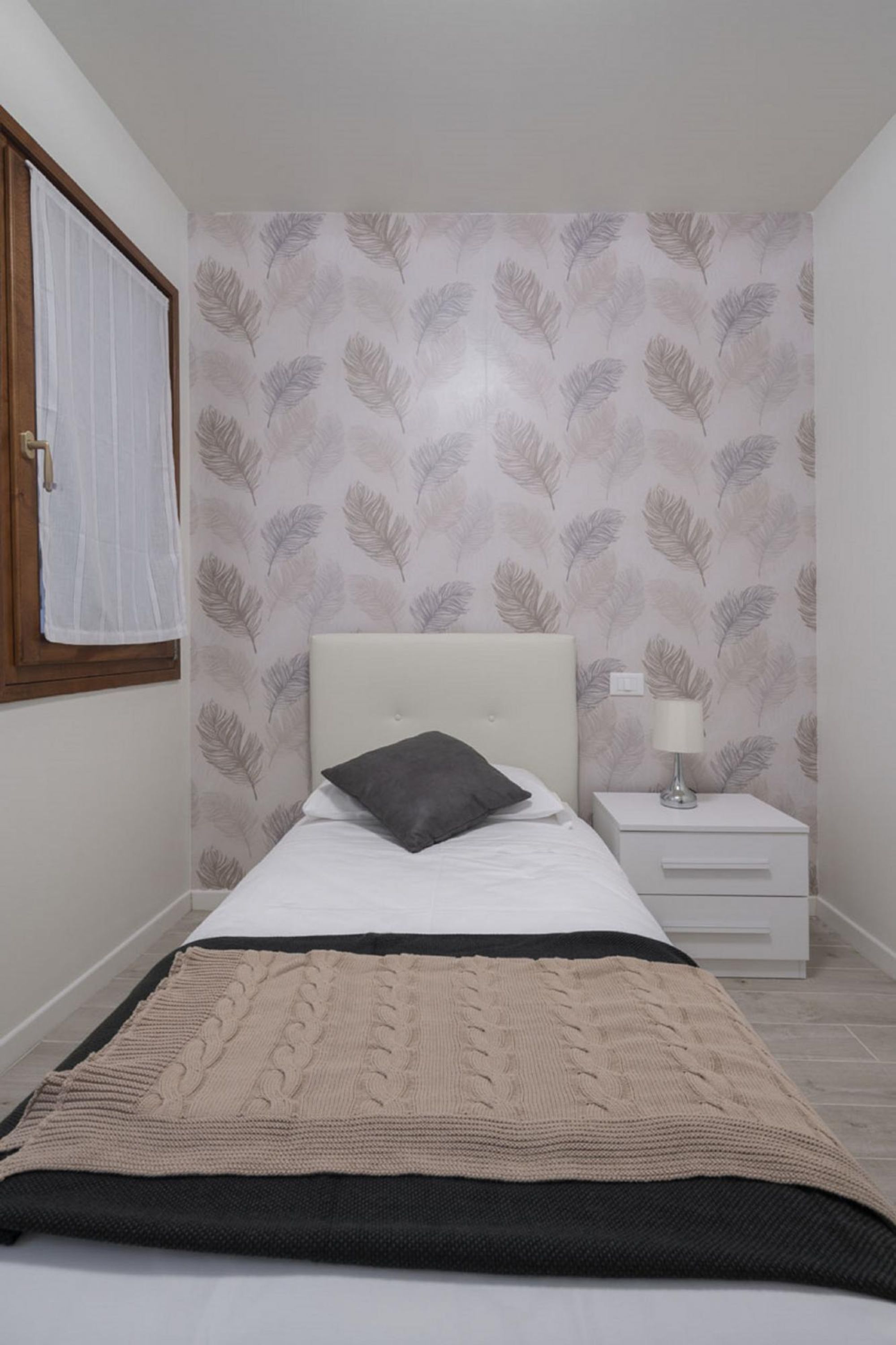 A bedroom with a bed, a nightstand, a lamp, a window with a curtain, and a wall with a leaf pattern.