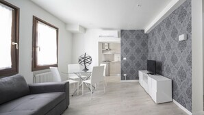 A modern living room with a grey sofa, a glass dining table with white chairs, a white TV stand, and patterned wallpaper. - San Marco Style Canal View Apt 1 by Wonderful Italy (Venice)