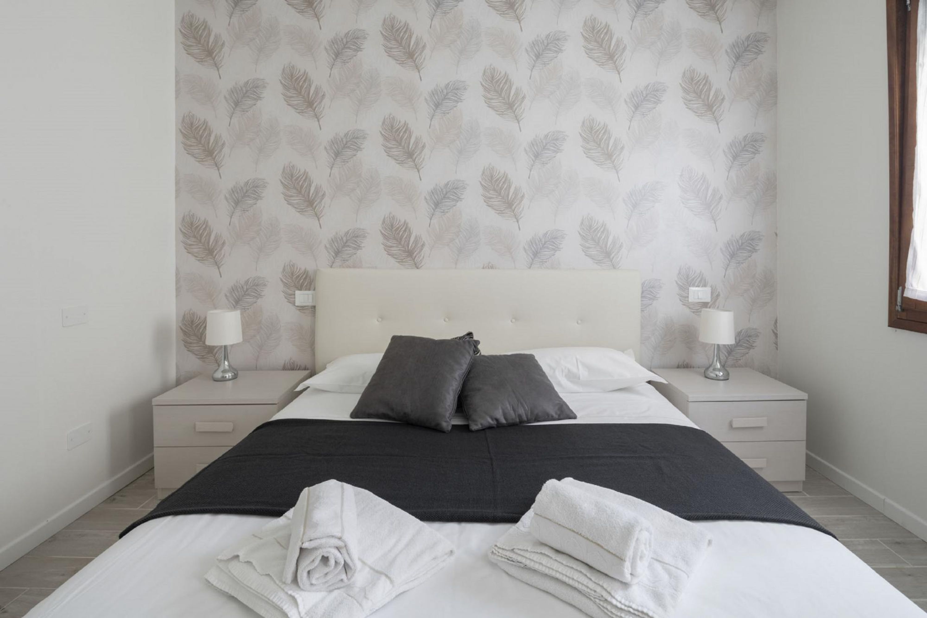 A neatly made bed with white linens and two pillows, flanked by bedside tables with lamps, in a room with a leaf-patterned wallpaper.