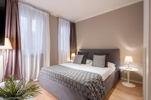 A bedroom with a bed, two bedside tables, a potted plant, and sheer curtains.