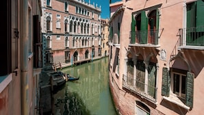 A narrow canal in a historic European city with buildings on either side, a gondola docked at the canal's edge, and a clear blue sky. - San Marco Suite 1 by Wonderful Italy (Venice)
