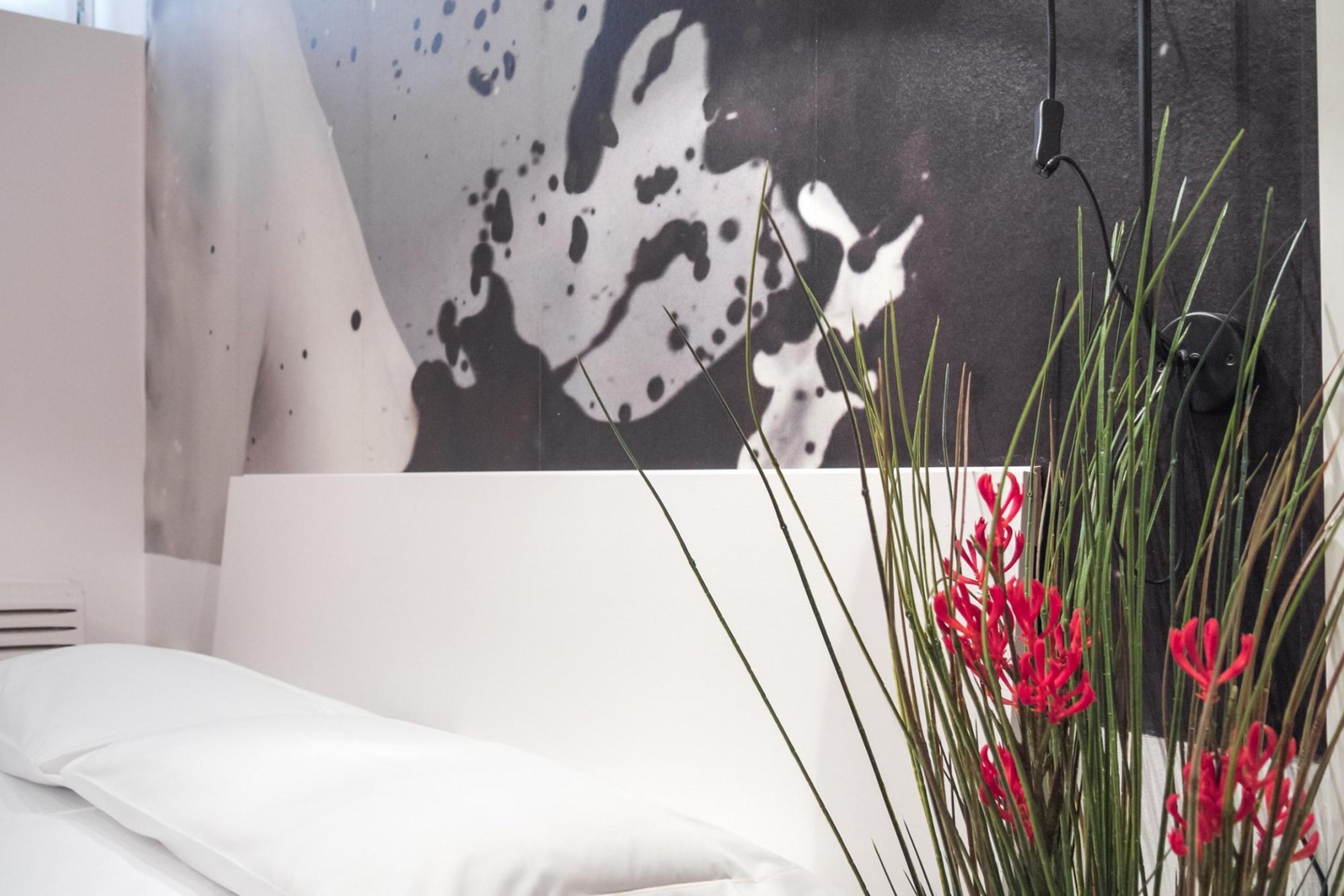 Una habitación moderna con una cama blanca, un cuadro abstracto grande y flores rojas en un jarrón.