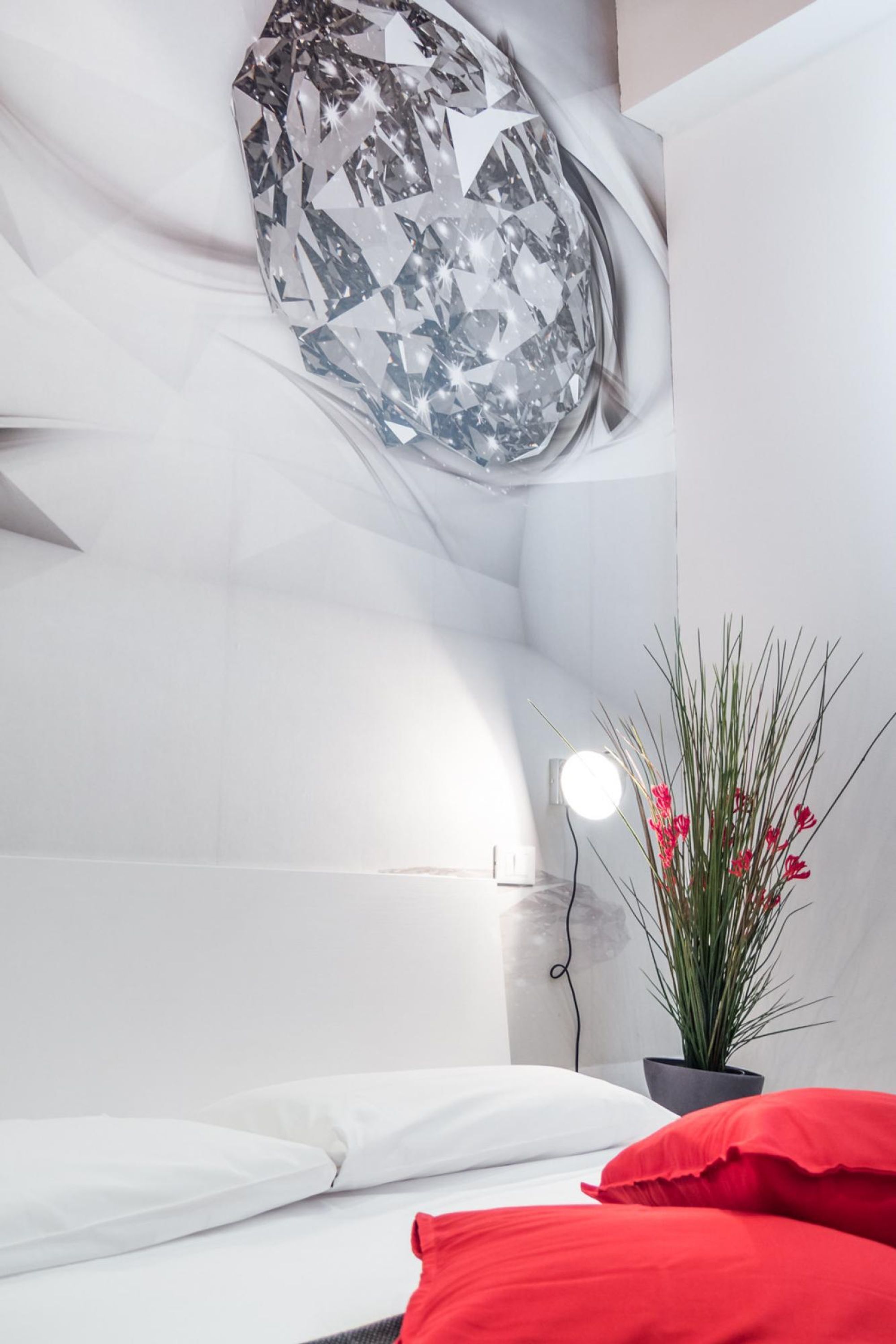 Ein Schlafzimmer mit einem Bett, einem roten Kissen, einer Topfpflanze und einer Wandleuchte.