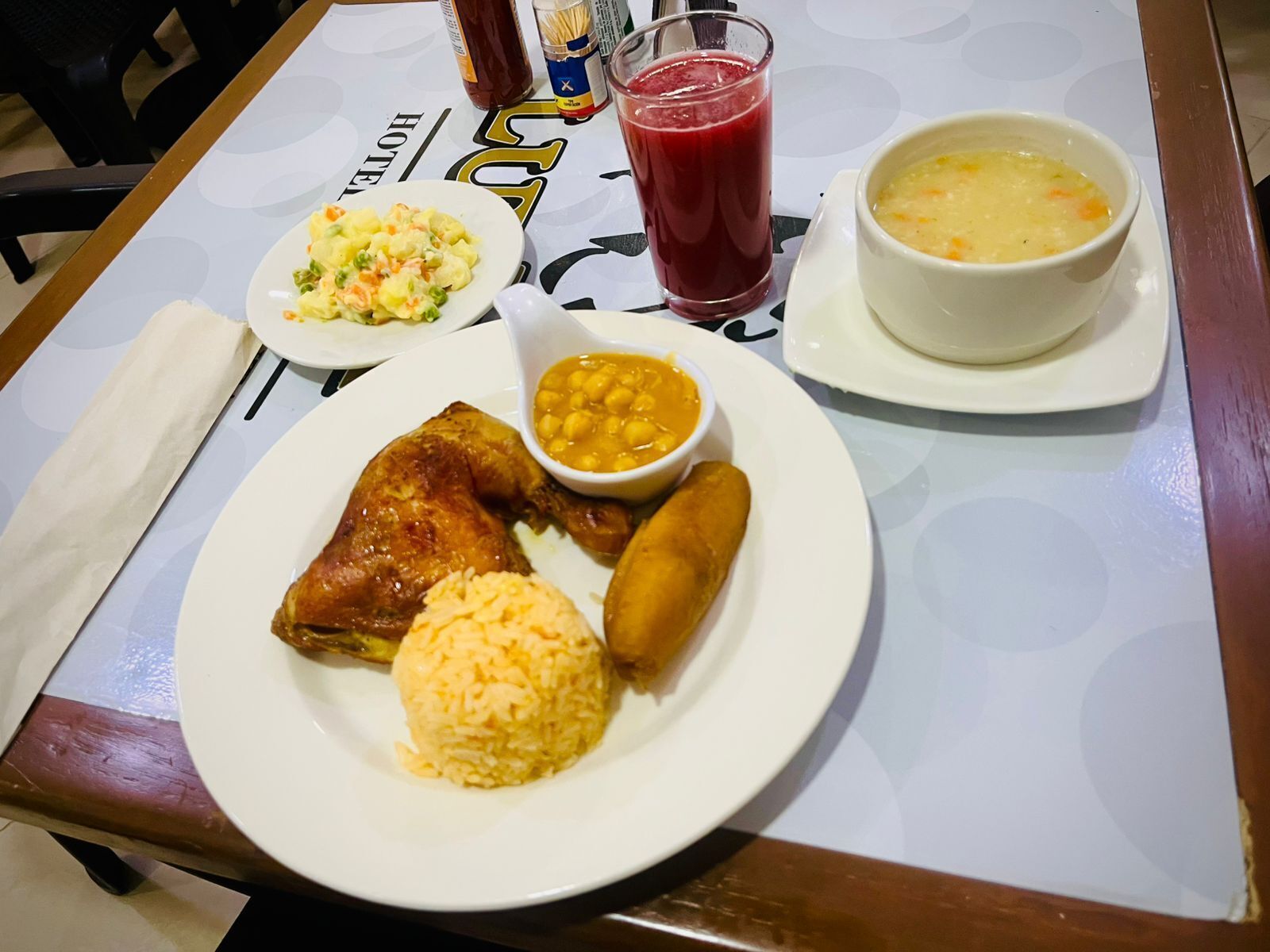A table with a meal consisting of rice, a fried item, a baked item, a bowl of soup, a bowl of chickpeas, and a glass of red drink.
