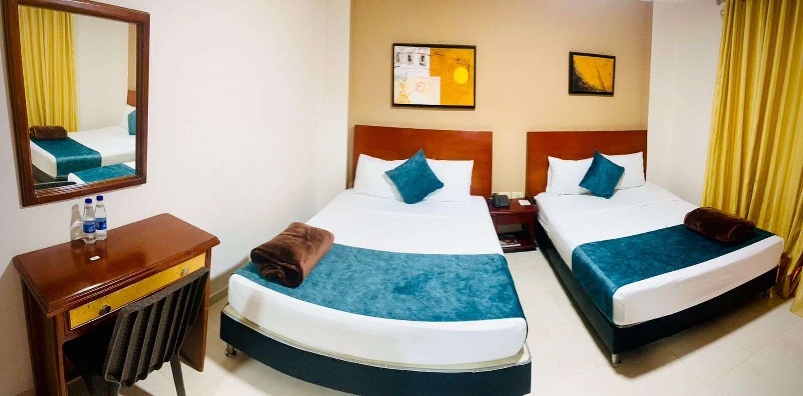 Habitación de hotel con dos camas, una mesita de noche con lámpara, un espejo y obras de arte en las paredes.