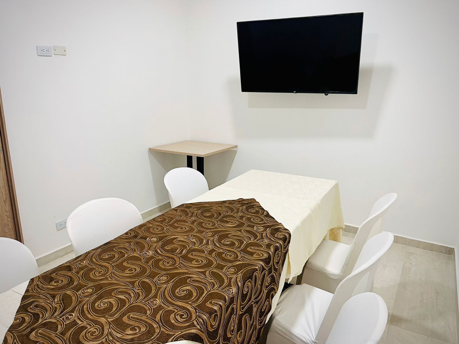 Une chambre d’hôtel avec un téléviseur fixé au mur, une petite table recouverte d’une nappe de couleur claire et un chemin de table à motifs.