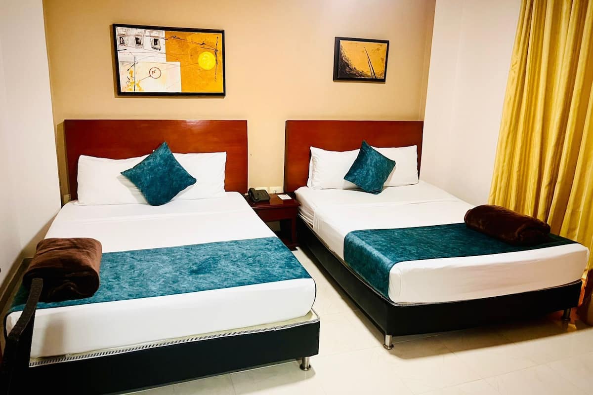 A hotel room with two beds, each with a wooden headboard, white bedding, and teal throw pillows. There are two framed artworks on the wall, a telephone on the nightstand, and a yellow curtain.