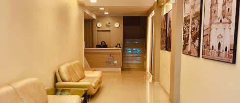 Zona de recepción de hotel con un mostrador, un área de estar con sofás y una escalera que sube a un piso superior.
