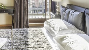 A hotel room with a bed, a nightstand, a lamp, a window with curtains, a framed picture on the wall, and a view of buildings outside. - San Marco Deluxe Canal View Balcony by Wonderful Italy (Venice)