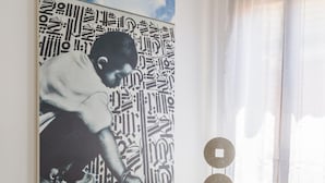 A room with a large abstract painting on the wall, a small table with a plant and a speaker, and a window with sheer curtains. - Accademia Charm 6 by Wonderful Italy (Venice)