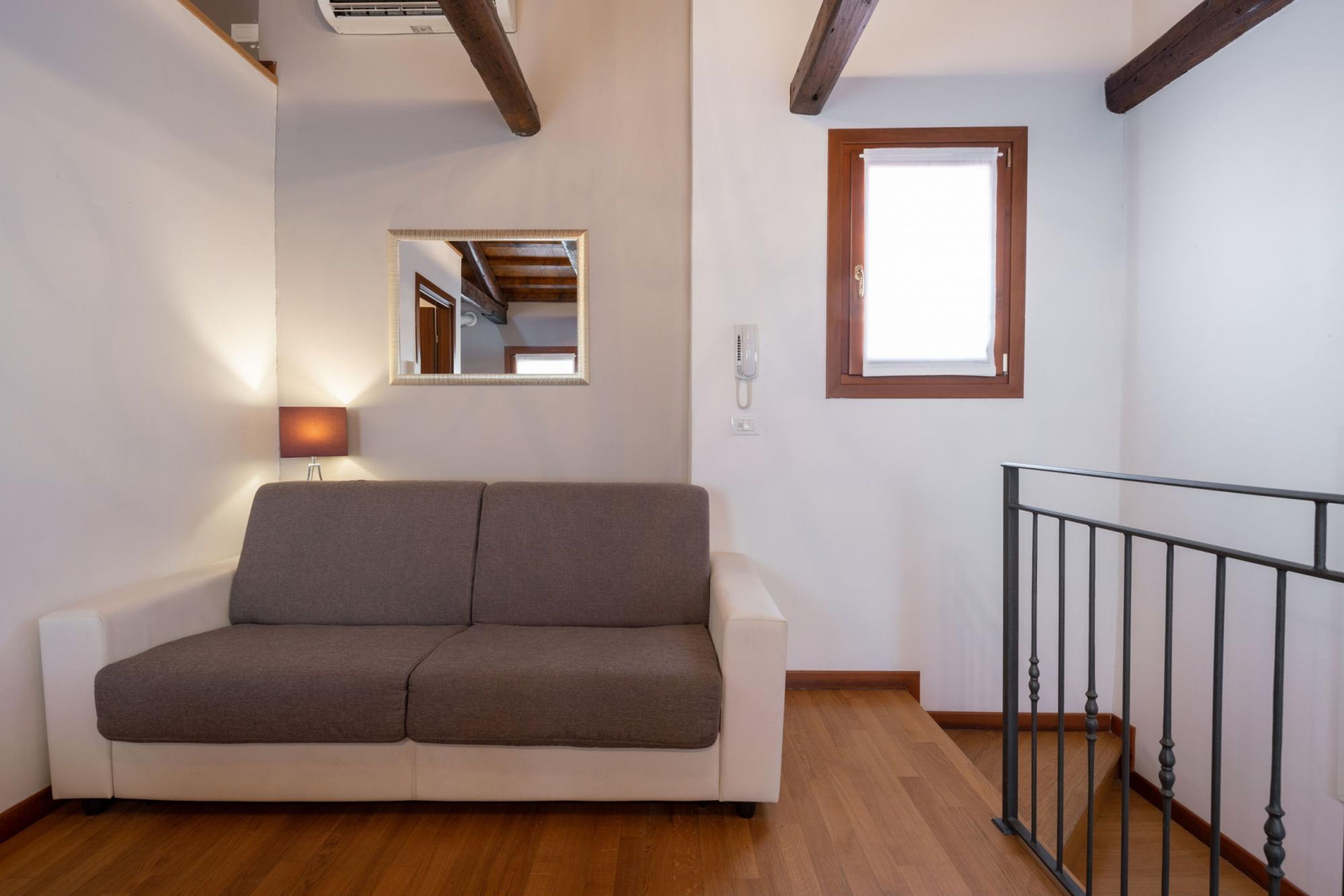 A modern living room with a sofa, a small window, a mirror, and a staircase with a railing.