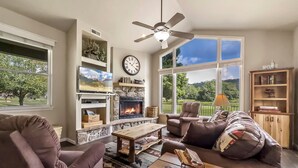 A living room with a fireplace, a television, a clock, and a view of the outdoors.