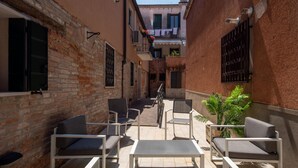 Um pátio estreito com mesas ao ar livre, um muro de tijolos e um edifício com varandas.