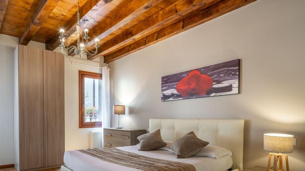 Quarto com cama grande, piso de madeira, lustre, mesa de cabeceira e janela com cortinas.