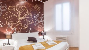Ein modernes Hotelzimmer mit einem großen Bett, einem Nachttisch mit Lampe, einer Wand mit floralem Muster und einem Fenster mit Jalousien.
