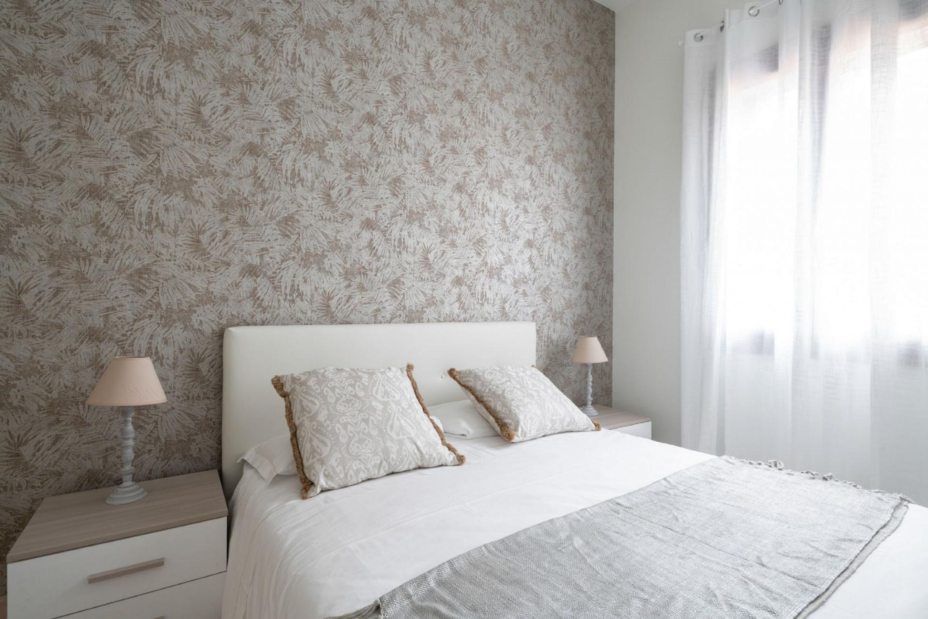 Une chambre avec un papier peint à motifs, un lit blanc agrémenté de deux coussins décoratifs, une table de chevet avec une lampe et une fenêtre ornée de rideaux légers.